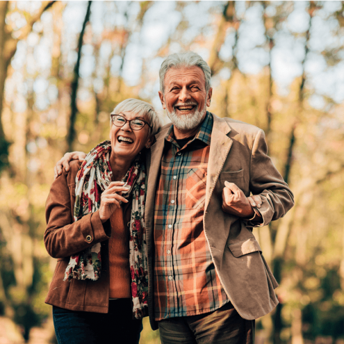 The image shows a joyful elderly couple standing together in an outdoor setting with trees in the background.