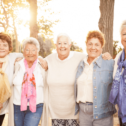 A group of five elderly women standing together outdoors in a park-like setting, smiling and wearing colorful scarves. They have their arms around each other and appear to be enjoying a sunny day.