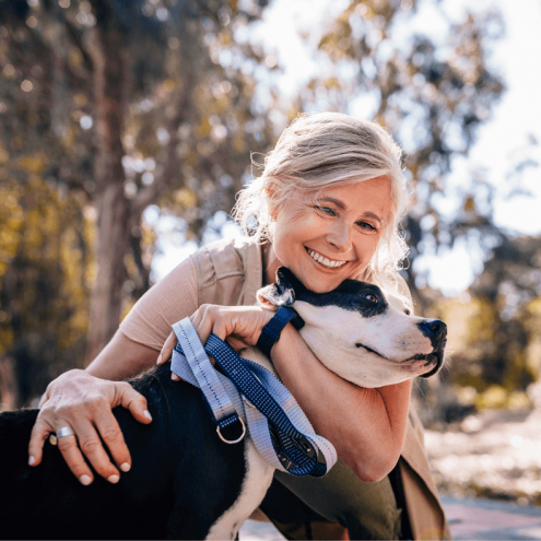 A senior woman smiling and hugging a black and white dog in a park. The dog is wearing a leash, and the woman appears joyful as she embraces the animal.