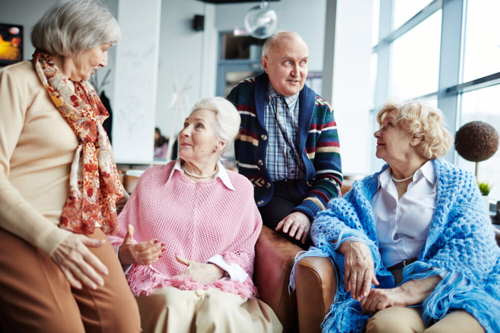 A group of elderly people sitting together in a cozy, well-lit room. Two women are standing, while two others are sitting on a sofa. They are all wearing colorful shawls and scarves, sharing a conversation.